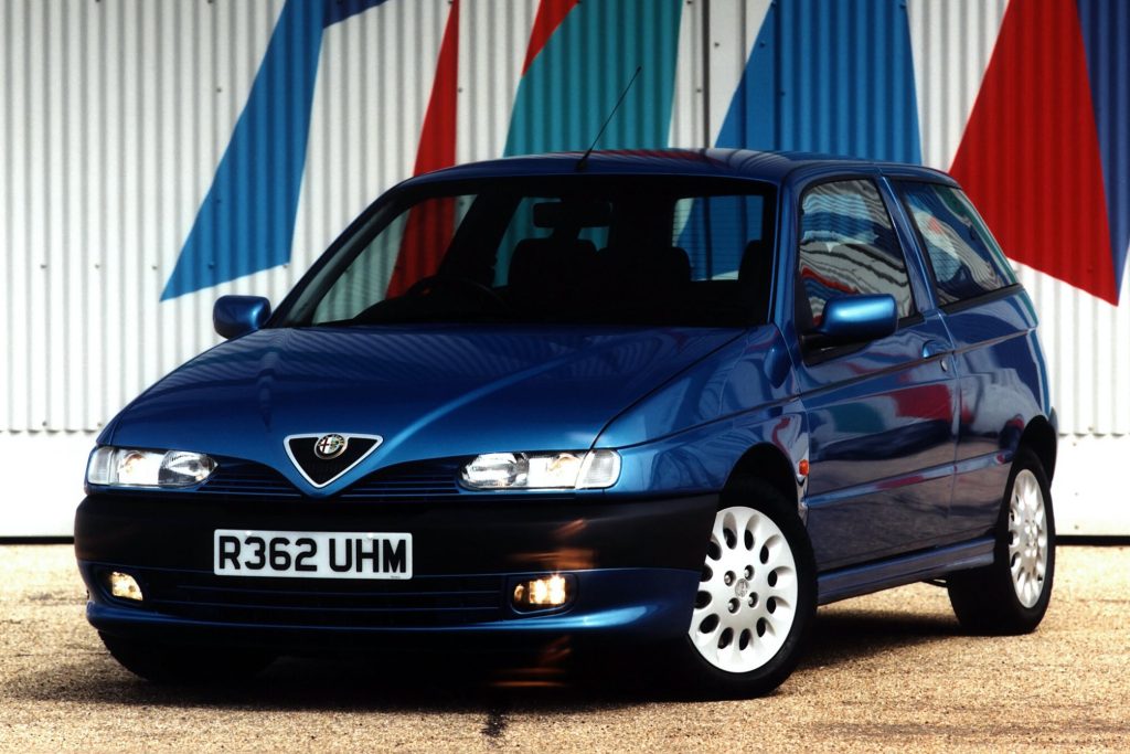 Blue Alfa Romeo 145 five-door hatchback parked in front of a colourful striped industrial wall with headlights on