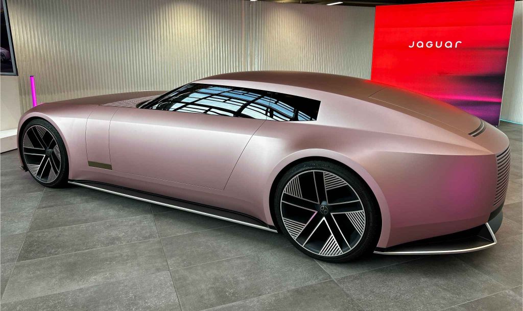 Rear three-quarter view of the Jaguar Type 00 concept car in a matte rose/blush pink finish, displayed indoors on grey tile flooring with a red Jaguar branded backdrop.