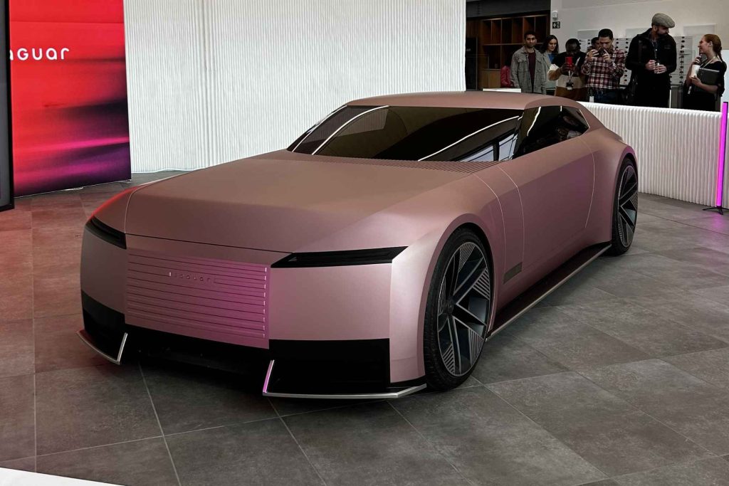 Front three-quarter view of the Jaguar Type 00 concept car in blush pink finish, shot from a lower angle showing the bold geometric front fascia with horizontal slat grille, flush lighting, and dramatic angular wheel arches, with onlookers visible in the background.
