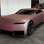 Front three-quarter view of the Jaguar Type 00 concept car in blush pink finish, shot from a lower angle showing the bold geometric front fascia with horizontal slat grille, flush lighting, and dramatic angular wheel arches, with onlookers visible in the background.