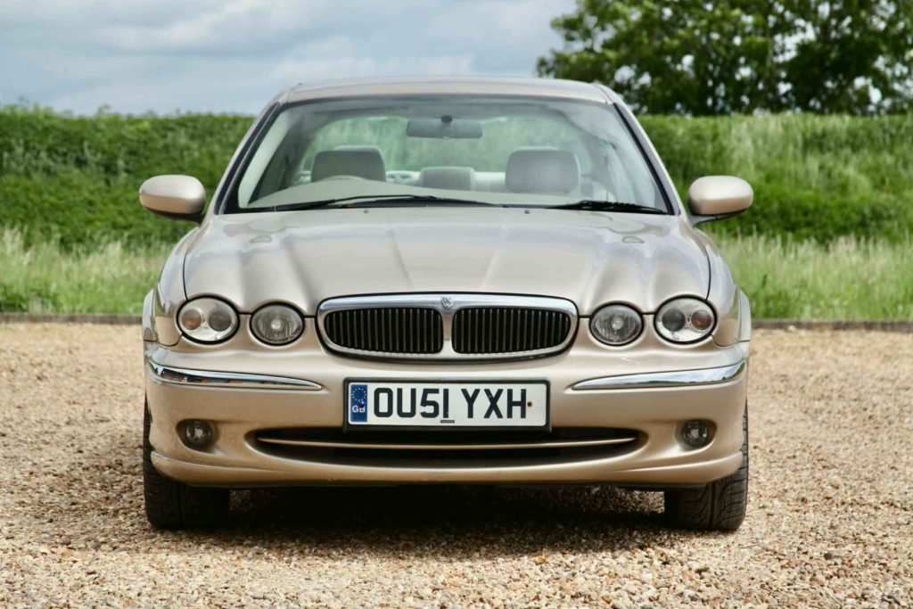 Head-on view of a Jaguar X-Type in gold metallic, parked on a gravel surface with green hedgerows and trees under a partly cloudy sky.
