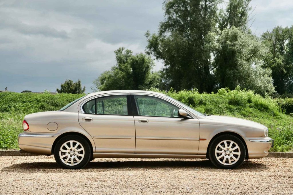 Side profile of a Jaguar X-Type in gold metallic, parked on a gravel surface with lush green hedgerows and an overcast sky in the background.