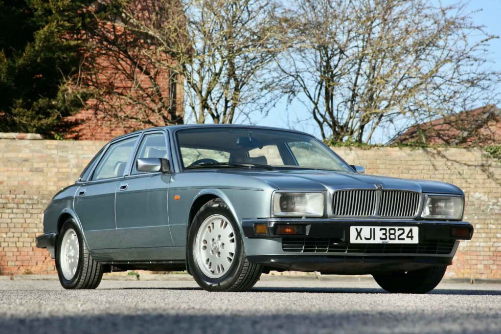 Front three-quarter view of a late-1980s Jaguar XJ6 Sovereign in steel blue metallic, photographed against a brick wall with bare winter trees.