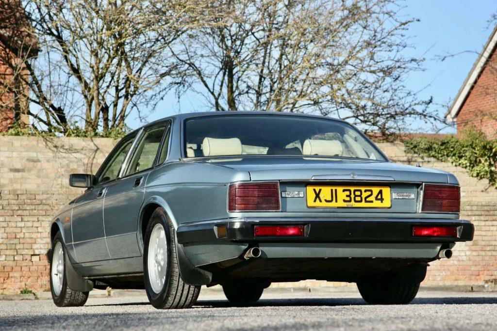 Rear three-quarter view of a Jaguar XJ6 Sovereign (registration XJI 3824) showing the wide rear lamp clusters, twin exhaust pipes, and traditional chrome badging, with the brick wall backdrop.