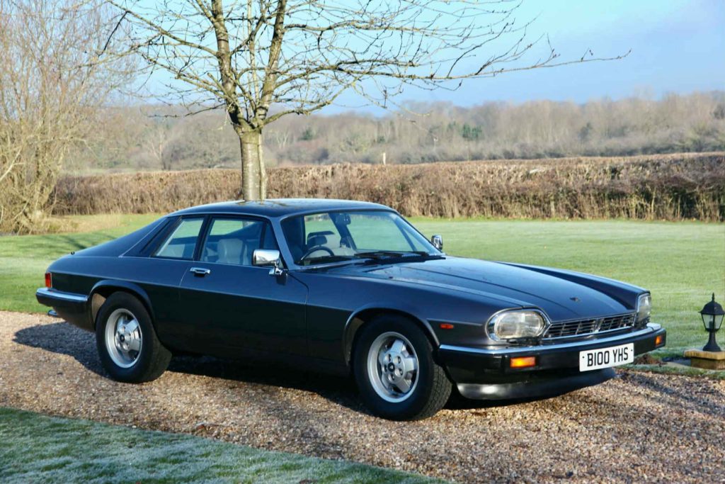Front three-quarter view of a Jaguar XJS HE coupe in dark navy/black, parked on gravel with a frosty winter rural landscape behind it.