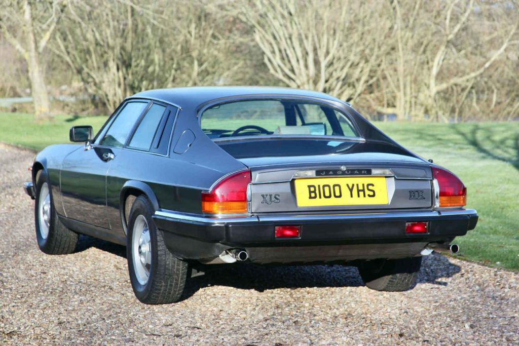 Rear three-quarter view of the same Jaguar XJS HE (registration B100 YHS) showing the wide rear deck, elongated tail lamps, twin exhausts, and "XJS HE" badging on the boot lid.