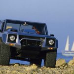 A silver-grey Lamborghini LM002 shot from a low front angle on a rocky coastal shoreline, with a driver visible inside wearing sunglasses. A white sailboat is visible on the blue sea in the background.
