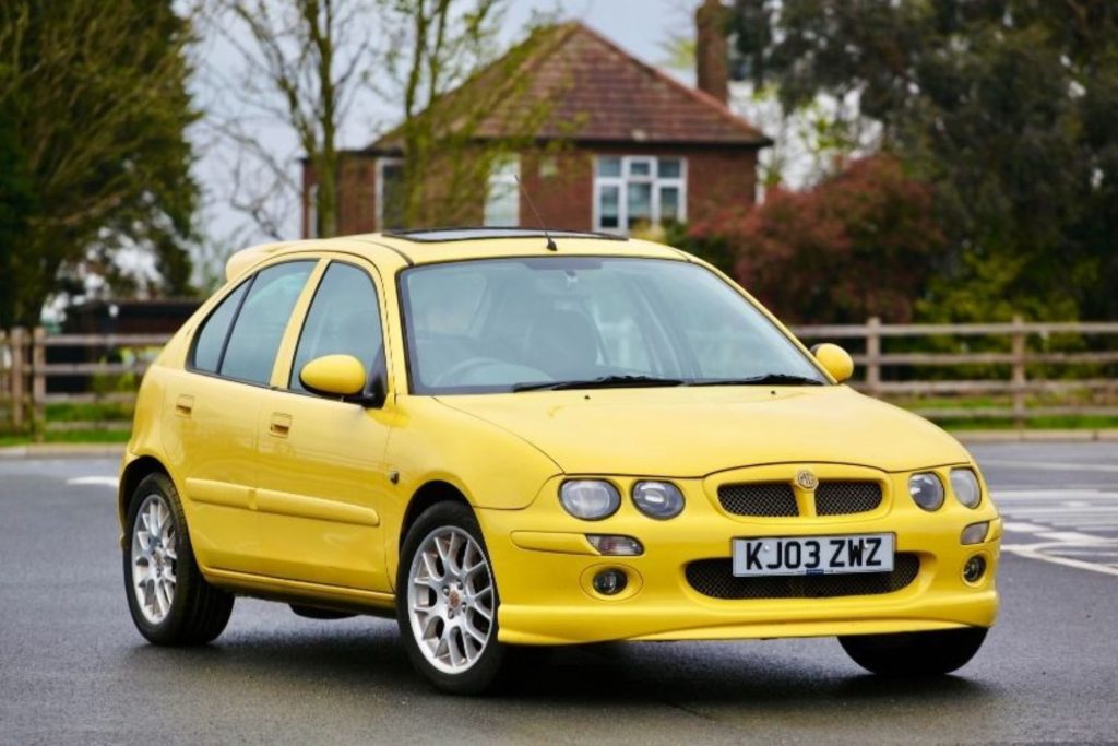 Yellow MG ZR five-door hatchback parked on a country road with a red brick house and trees in the background