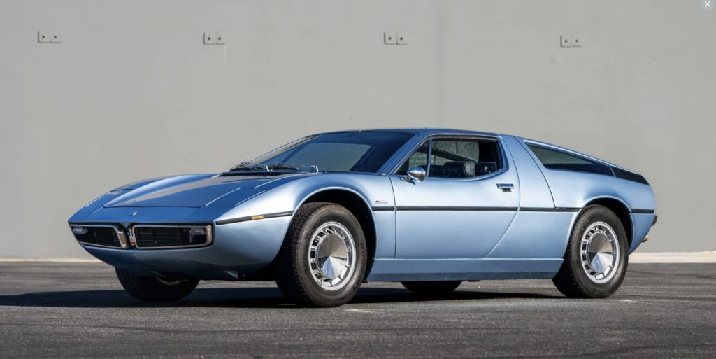 Front three-quarter view of a light blue Maserati Bora in bright sunlight, highlighting the clean bodywork, hood line, dark rear greenhouse louvers, and cone-hub alloy wheels.
