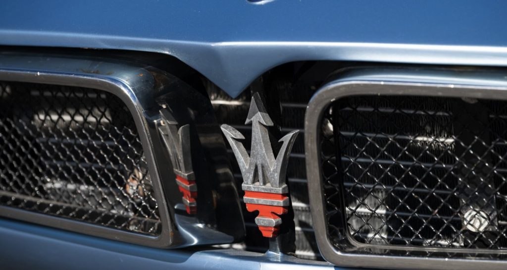 Close-up of the Maserati trident badge mounted in the twin-nostril front grille of a Bora, finished in aged silver with a red base, set against mesh grille inserts.