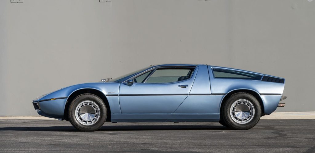 Full left-side profile of a light blue Maserati Bora parked against a grey wall, showcasing its low, wedge-shaped silhouette, fastback roofline, and period alloy wheels.