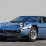 Front three-quarter view of a light blue Maserati Bora showing its low nose, twin round headlights, divided front grille with trident badge, and sweeping hood with central power bulge.