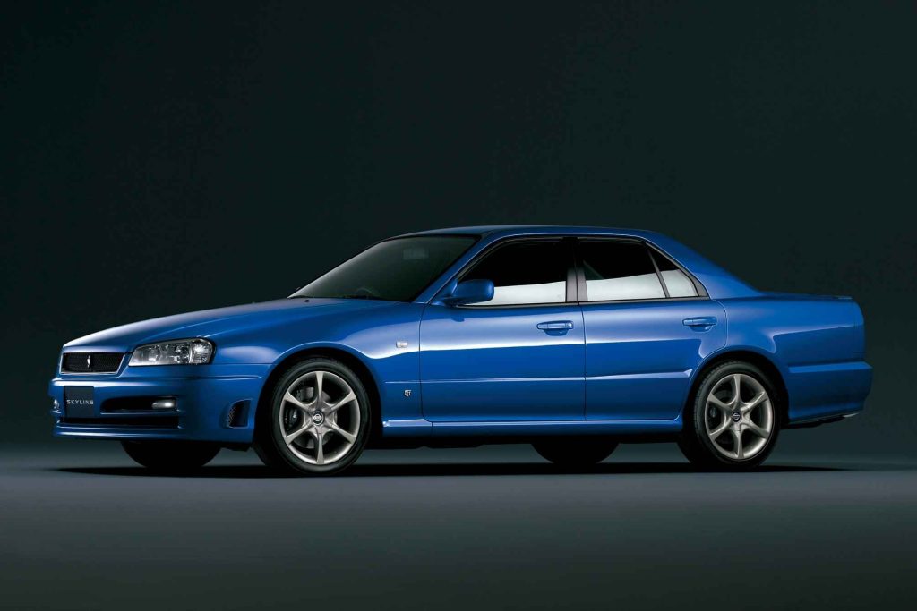 Bayside Blue Nissan Skyline R34 four-door sedan photographed from the front three-quarter angle in a studio, displaying the full profile with sport alloy wheels and body kit