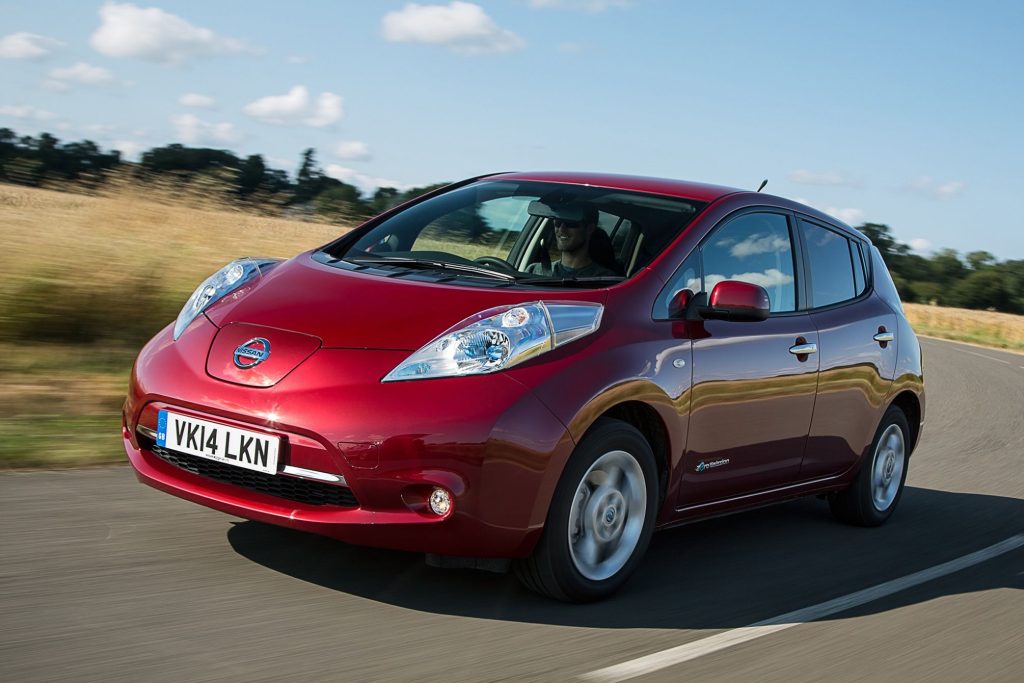 Red Nissan Leaf first-generation electric hatchback driving on a rural road in summer, front three-quarter view