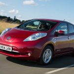 Red Nissan Leaf first-generation electric hatchback driving on a rural road in summer, front three-quarter view
