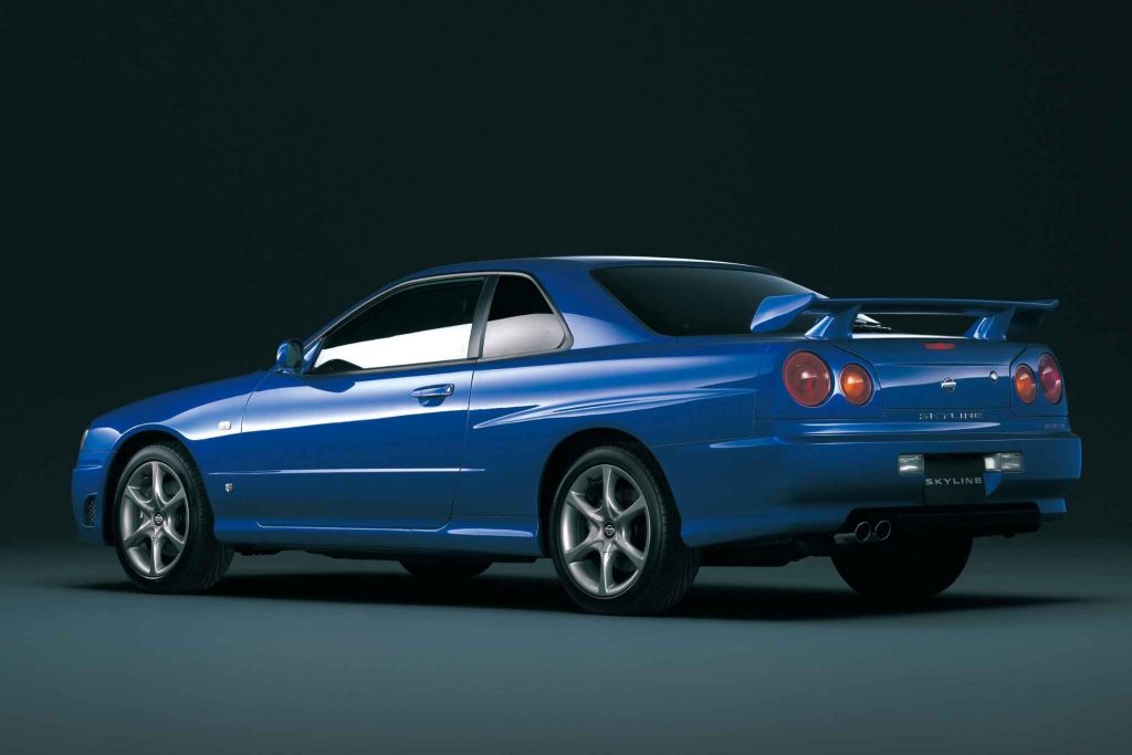 Bayside Blue Nissan Skyline R34 GT coupe photographed from the rear three-quarter angle in a studio, showing the iconic round taillights, rear spoiler, and dual exhaust