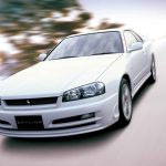 White Nissan Skyline R34 photographed from the front three-quarter angle in motion on a road, with motion blur in the background emphasizing speed