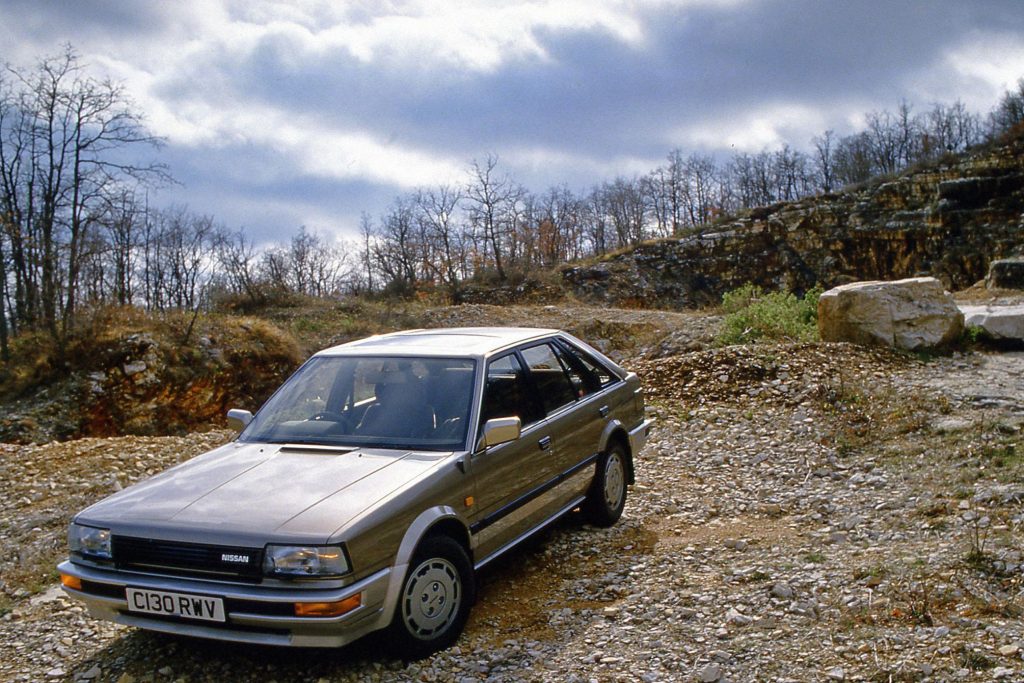 Gold Nissan Bluebird T12 four-door saloon parked on a rocky gravel track with bare trees and dramatic cloudy sky, UK
