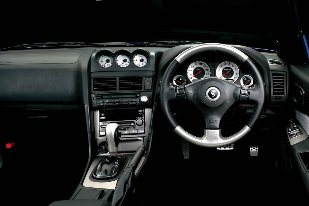 Right-hand drive cockpit of a Nissan Skyline R34 showing the three-gauge cluster on the dashboard, sport steering wheel with Skyline emblem, automatic transmission shifter, and white-faced instrument cluster
