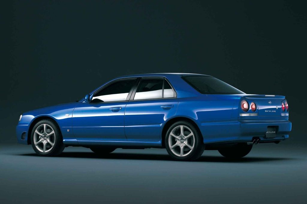 Bayside Blue Nissan Skyline R34 four-door sedan photographed from the rear three-quarter angle in a studio, showing the round taillights and subtle trunk spoiler