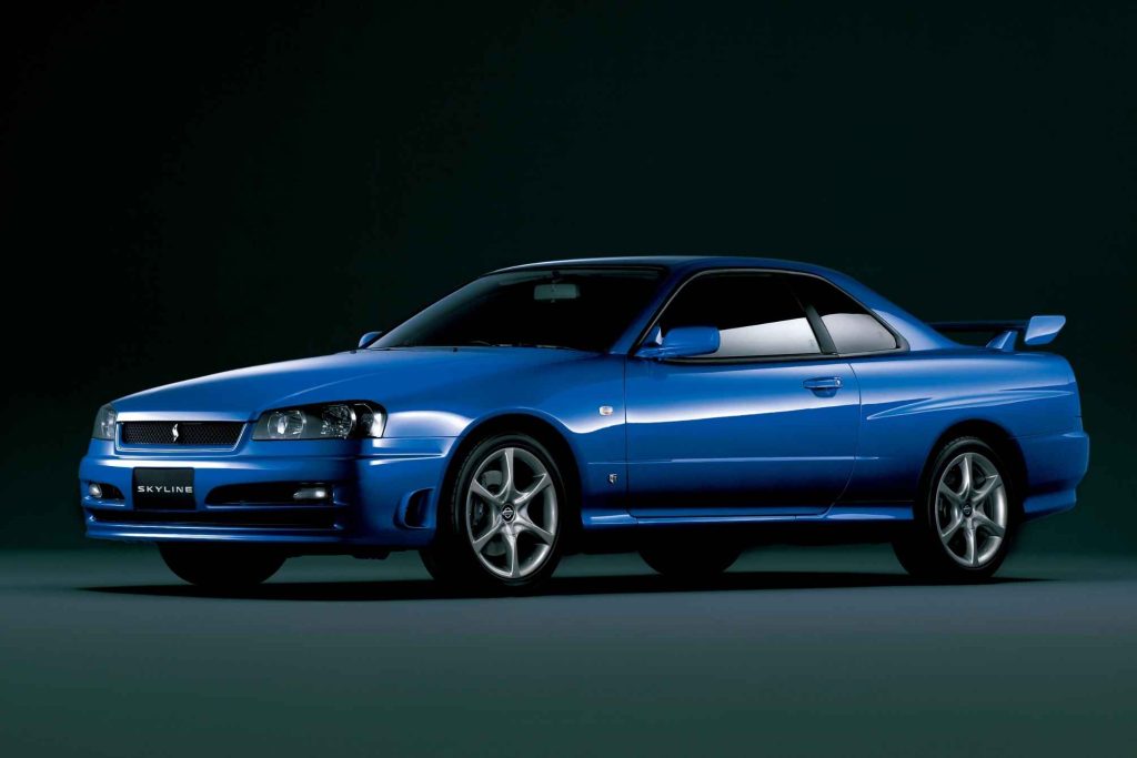 Bayside Blue Nissan Skyline R34 GT coupe photographed from the front three-quarter angle in a studio, showing the mesh grille, projector headlights, front lip spoiler, and rear wing