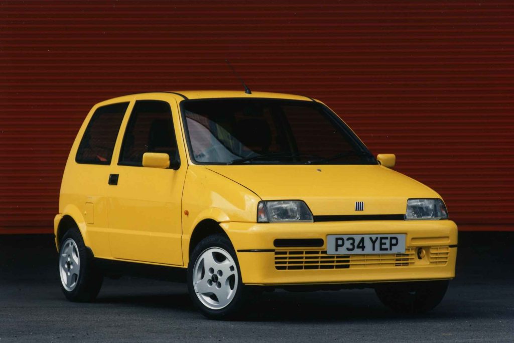Yellow Fiat Cinquecento Sporting hatchback photographed three-quarter front view against a red corrugated metal background