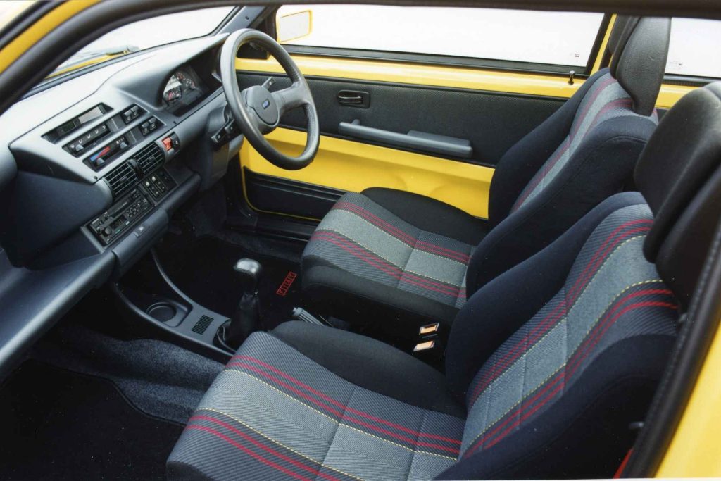 Interior of a yellow Fiat Cinquecento showing the dashboard, steering wheel, and grey cloth seats with red and yellow striped upholstery