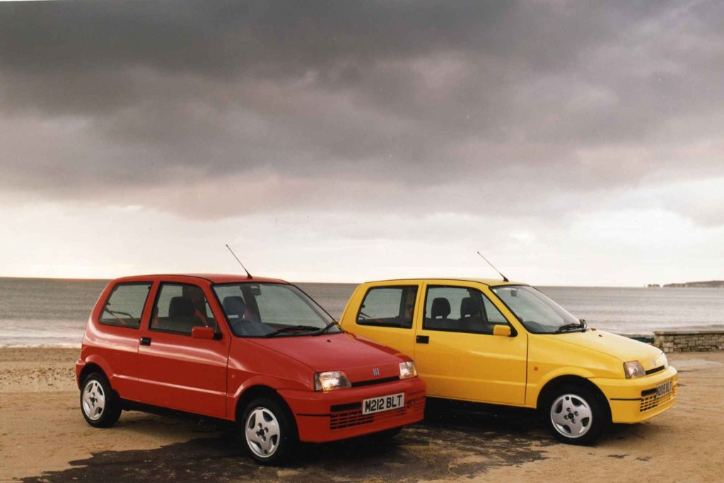 Red and yellow Fiat Cinquecento Sporting models parked side by side on a beach under a dramatic cloudy sky