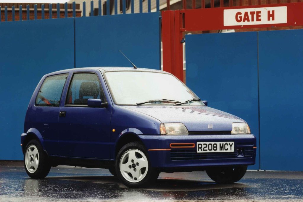 Blue Fiat Cinquecento Sporting front three-quarter view in the rain outside a blue industrial building with a Gate H sign