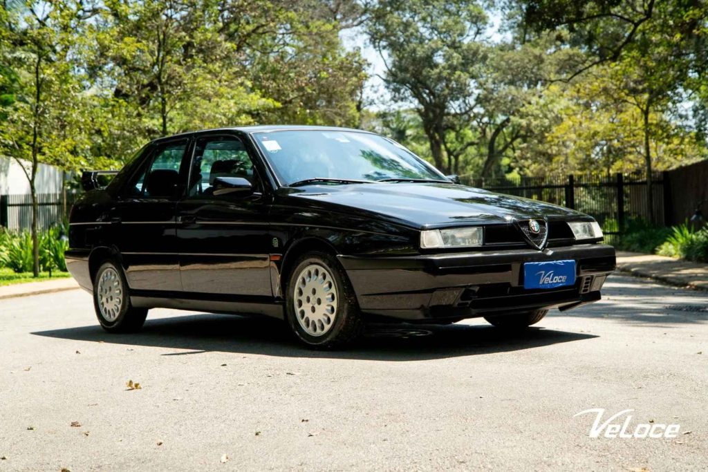 A black Alfa Romeo 155 sedan parked on a driveway surrounded by trees, showing the front three-quarter view with multi-spoke alloy wheels, photographed for a dealer called Veloce.