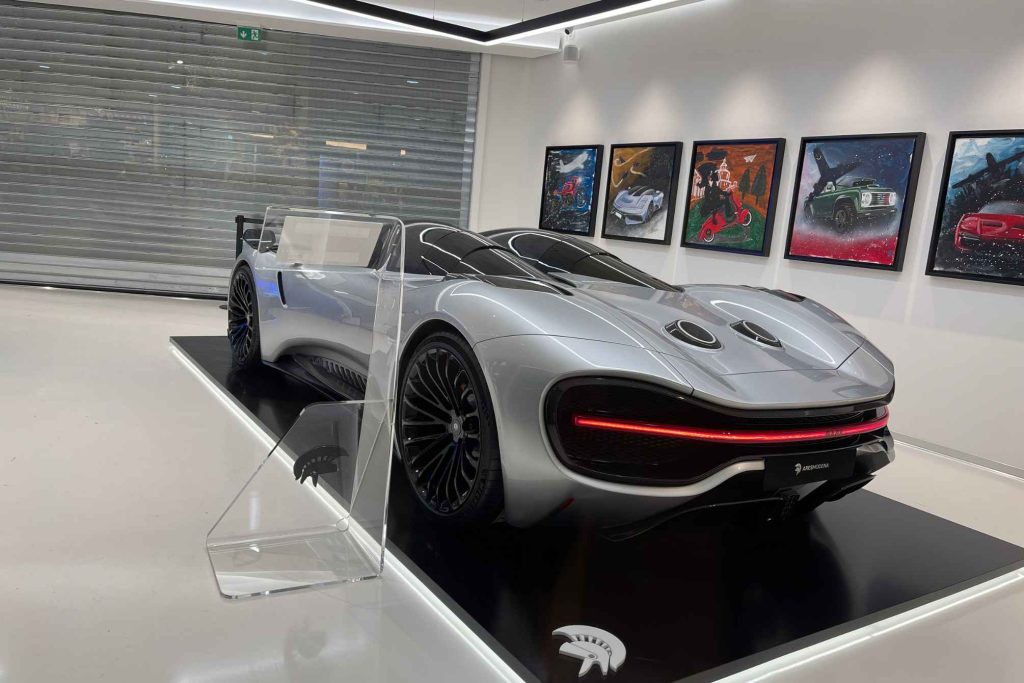 A silver Ares Modena concept hypercar on a black display plinth, with colorful automotive artworks lining the gallery wall behind it.