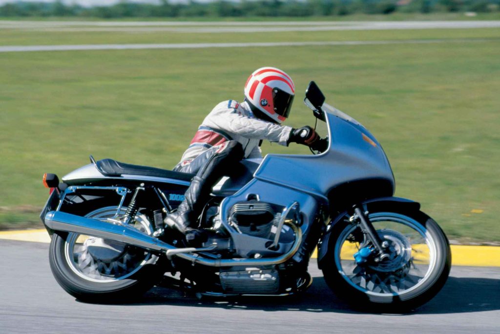 BMW R100RS in action on a runway or test track, rider tucked behind the wind-tunnel-developed full fairing, showing the landmark sports-tourer in profile