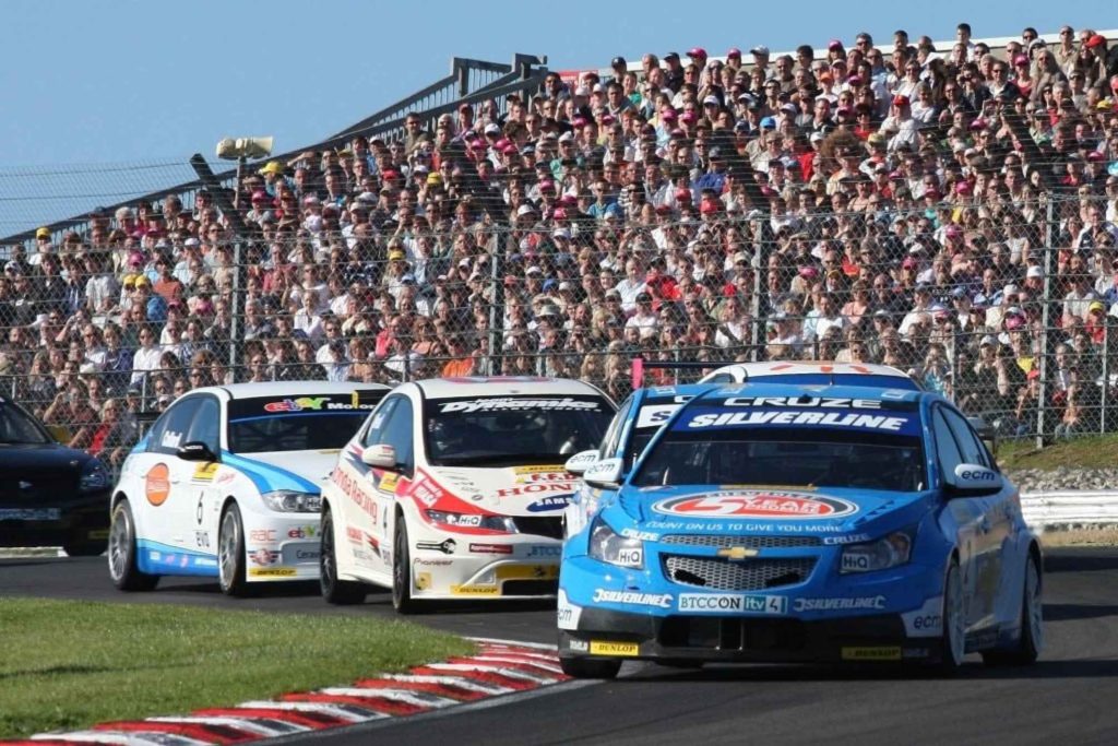 A British Touring Car Championship (BTCC) race at a packed circuit, led by a blue Chevrolet Cruze (#1, Silverline-sponsored), followed by a white Honda and other competitors, with a large crowd filling the grandstands behind a safety fence.