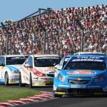A British Touring Car Championship (BTCC) race at a packed circuit, led by a blue Chevrolet Cruze (#1, Silverline-sponsored), followed by a white Honda and other competitors, with a large crowd filling the grandstands behind a safety fence.