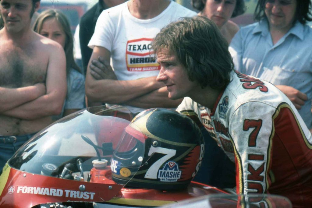 Barry Sheene in close-up beside his number 7 Texaco Heron Team Suzuki machine, wearing leathers and looking relaxed in a paddock crowd