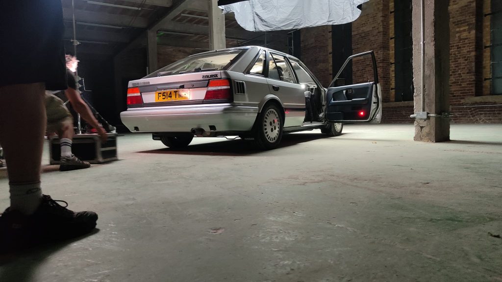 The Nissan Bluebird lit by professional studio lights inside a large industrial brick warehouse, door open, during a film or photo shoot.