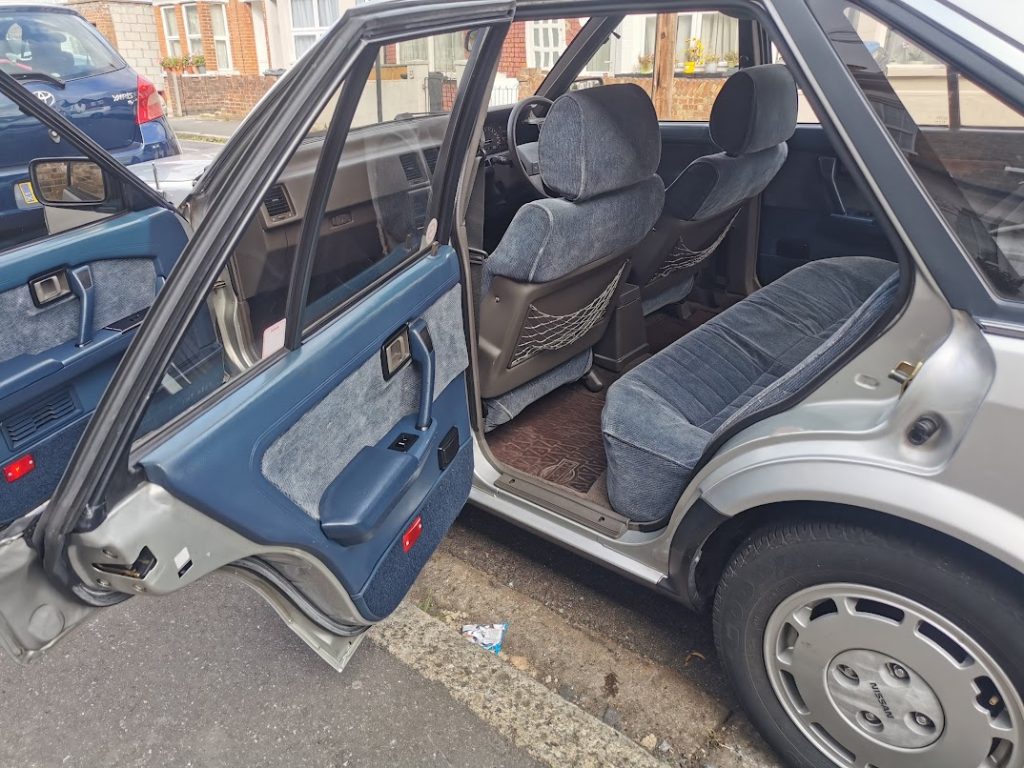 Interior view of the Nissan Bluebird with both rear doors open, showing blue-grey ribbed cloth seats, door cards, and patterned floor mats.
