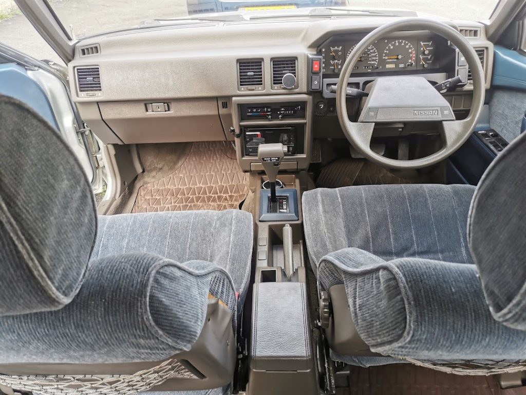 Dashboard and front cabin interior of the Nissan Bluebird, showing the analogue instrument cluster, automatic gear selector, centre console, and blue-grey cloth seats.
