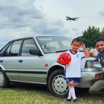 A man and a young boy in an England football kit holding a red ball pose in front of the silver Nissan Bluebird on open ground, with a British Airways aircraft passing overhead.
