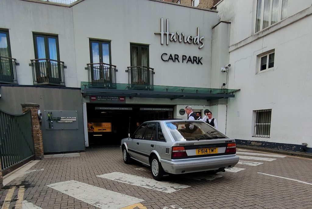 The silver Nissan Bluebird (F514 TMT) pulling out of the Harrods Car Park entrance in Knightsbridge, with uniformed Harrods staff watching nearby.