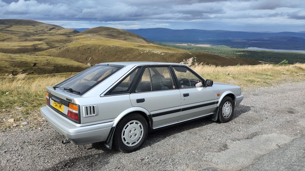 A silver Nissan Bluebird hatchback parked on a gravel road overlooking a sweeping Scottish Highland landscape with rolling hills, pine forests, and a loch in the distance under a dramatic cloudy sky.