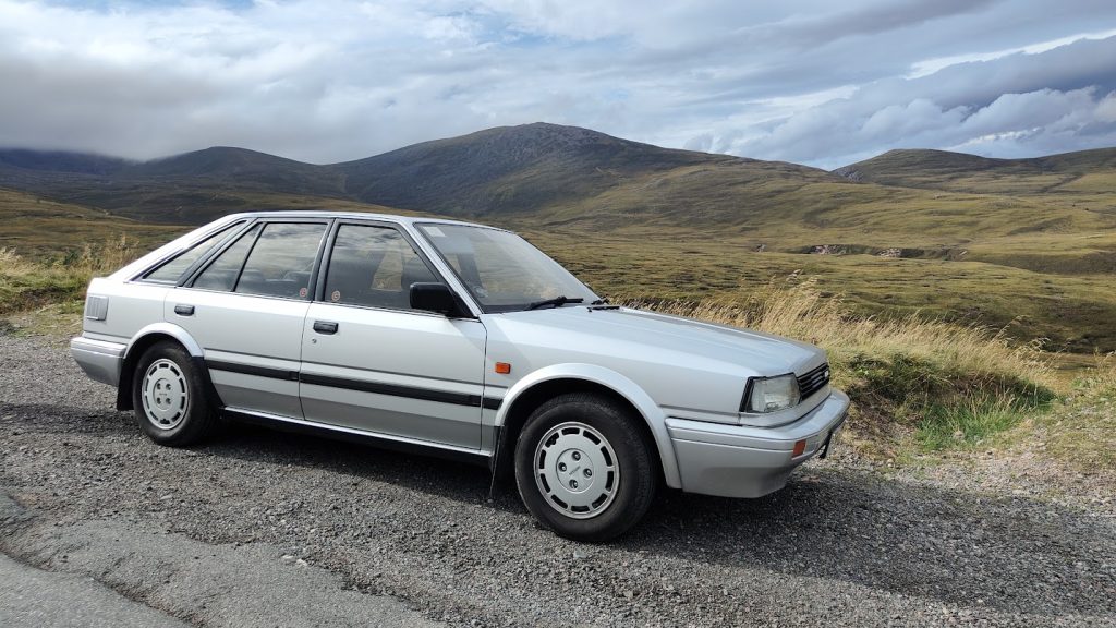 Side profile of a silver Nissan Bluebird parked on a remote Highland road, with rugged moorland hills and overcast skies behind it.
