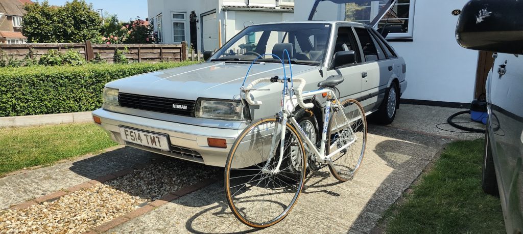 Front view of a silver Nissan Bluebird (registration F514 TMT) parked on a driveway with a vintage white Raleigh road bike leaning against it.