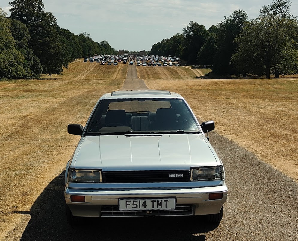 Head-on view of the silver Nissan Bluebird on a long straight avenue lined with trees, with a large gathering of classic cars visible in the far distance.