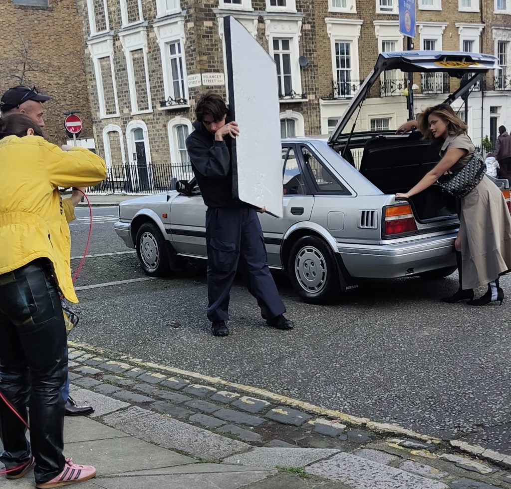 Behind-the-scenes street photo shoot in London, with a model posing at the open boot of the silver Nissan Bluebird while a crew member holds a reflector panel.