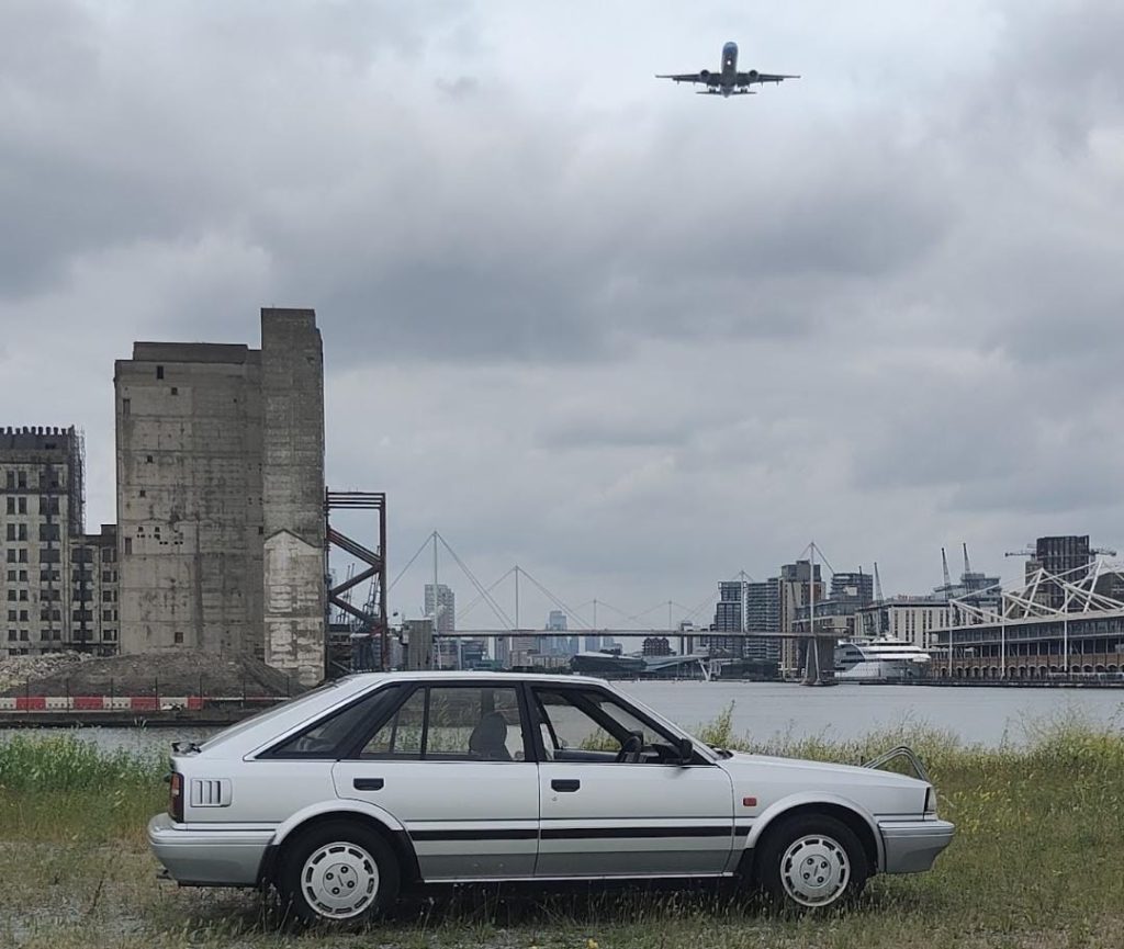 Side profile of the Nissan Bluebird parked on rough ground beside a river with a post-industrial London skyline and a passenger jet flying overhead.
