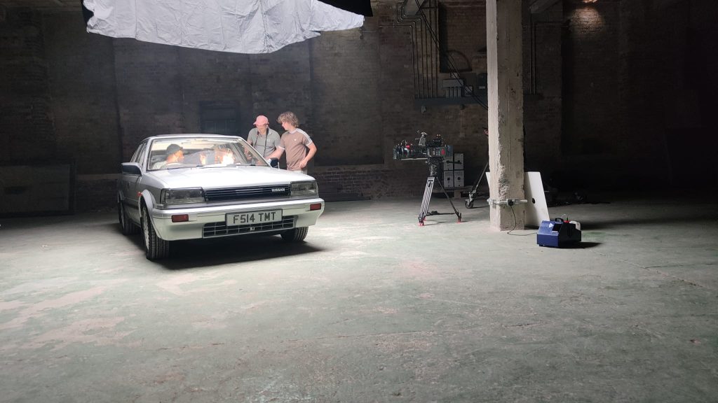 Film crew setting up shots around the front of the Nissan Bluebird inside a dramatic brick warehouse studio space with professional camera equipment visible.