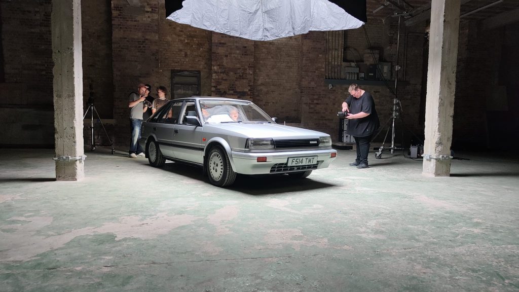 Wide shot of the Nissan Bluebird surrounded by a film crew in a large industrial warehouse, with an overhead diffusion silk and multiple photographers at work.