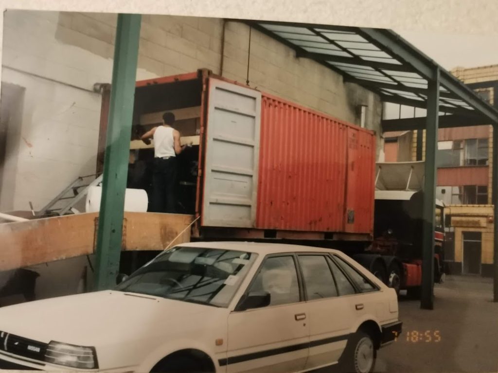 Vintage snapshot (timestamped 18:55) of a white Nissan Bluebird parked in an industrial yard beside a red shipping container being unloaded.