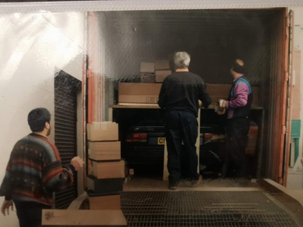 Vintage photo of three people unloading cardboard boxes from the back of a van or truck, with what appears to be a dark Nissan Bluebird visible inside.